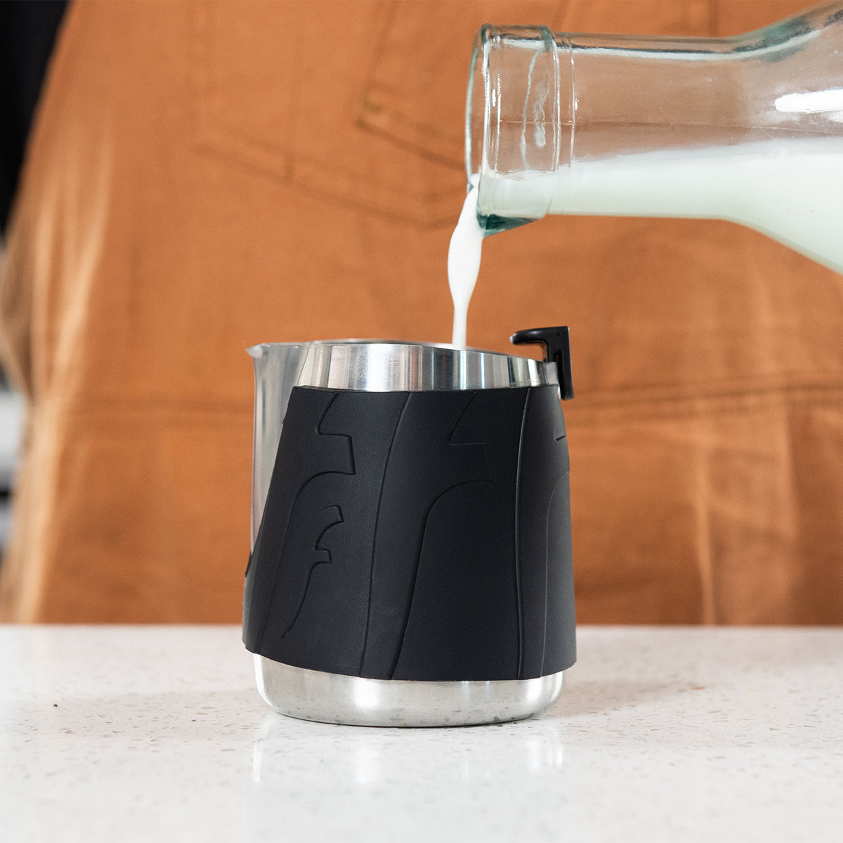Glass container pouring milk into a black and silver pitcher on a wooden surface.