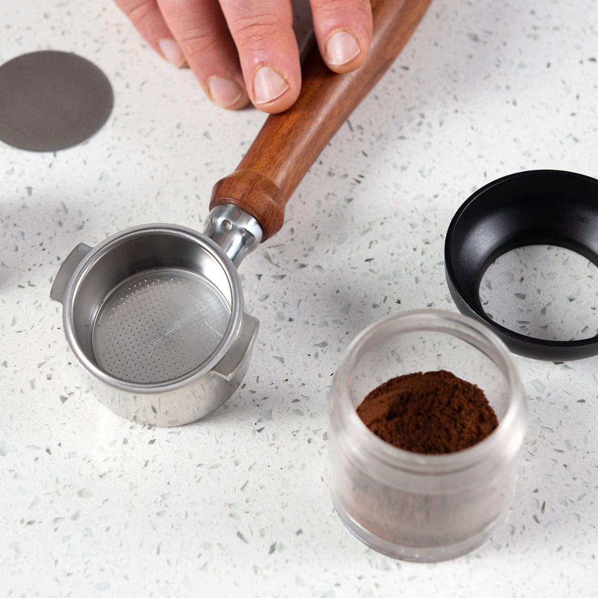 Hand holding a wooden-handled 49mm portafilter over a container of ground coffee on a light surface.