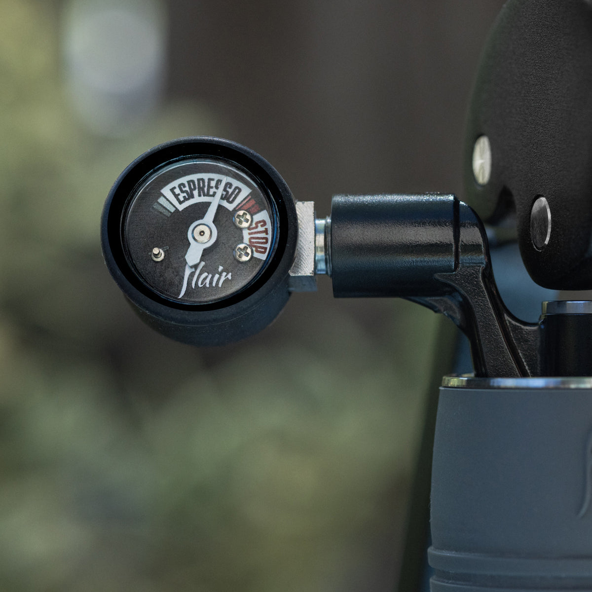 Close-up of a black espresso flow control valve with a circular dial labeled 'ESPRESSO' and 'FAIR', attached to a dark metal component against a blurred outdoor background.