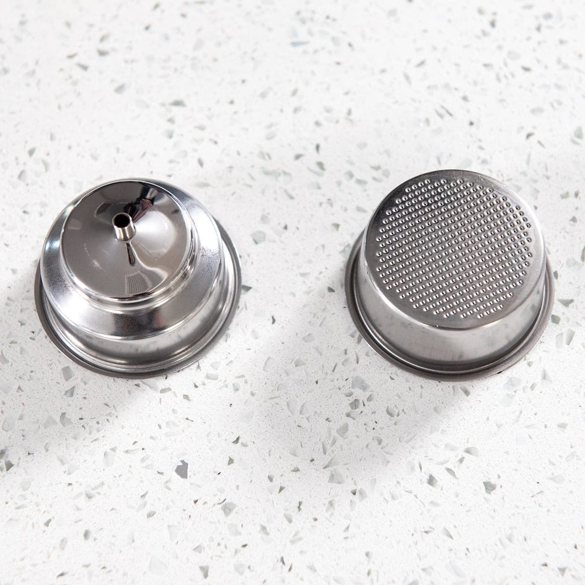 Two portafilter baskets on a white kitchen surface, one pressurized, the other bottomless.